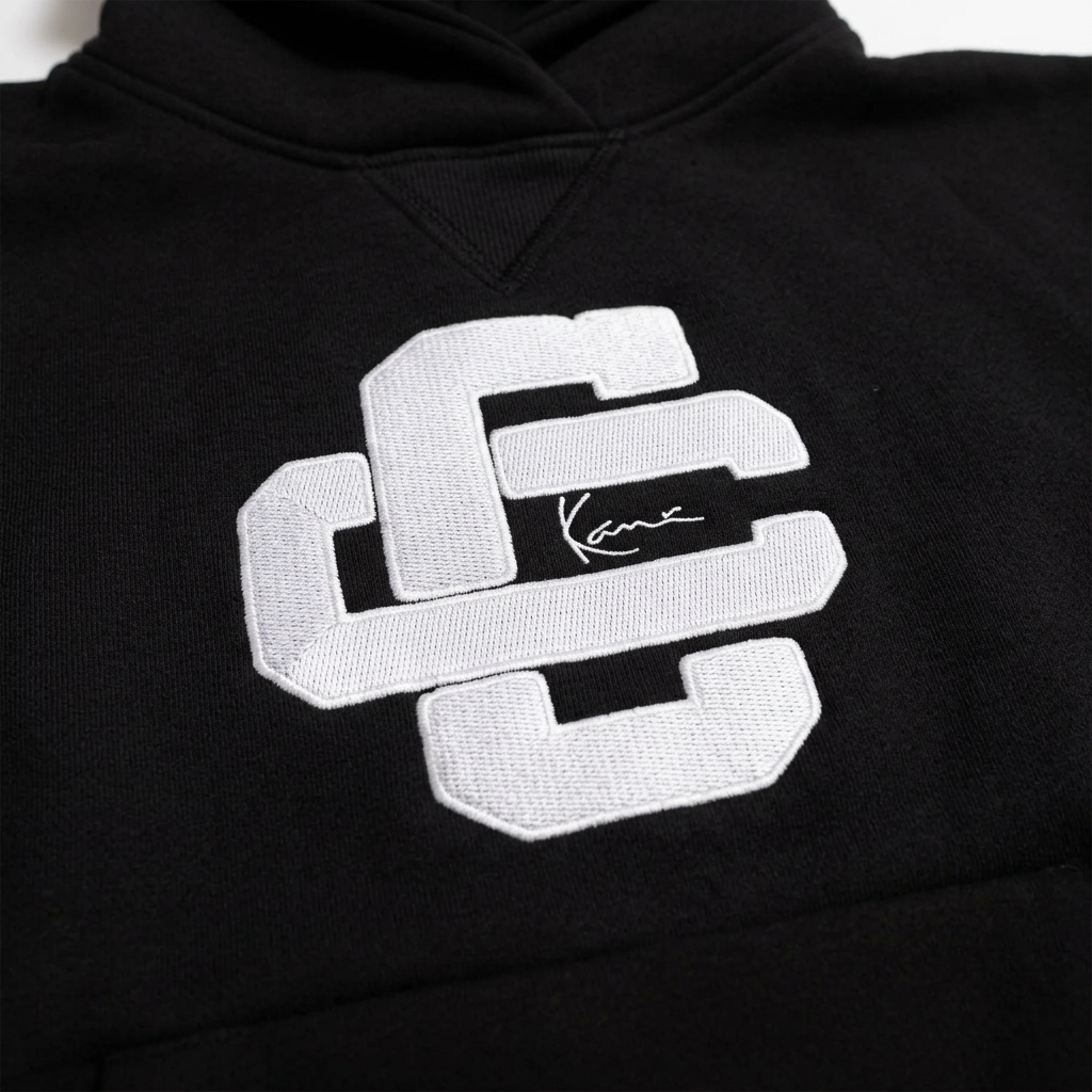 Close-up of the white Culture Creators x Karl Kani interlocking CC logo with Karl Kani signature on the chest of the black oversized hoodie