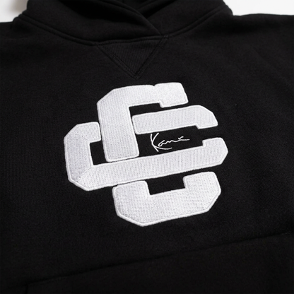 Close-up of the white Culture Creators x Karl Kani interlocking CC logo with Karl Kani signature on the chest of the black oversized hoodie